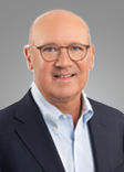 Richard Bane wears a blue colored shirt and jacket and smiles at the camera