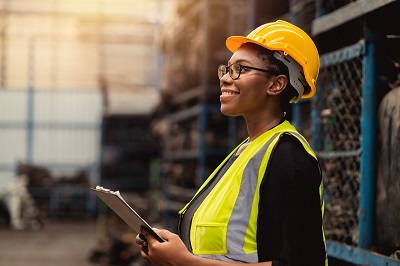 A woman wears a hardhat and safety vest and holds a clipboard inside of an industrial warehouse