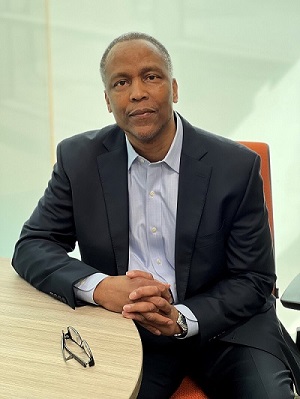 Anthony Samuels sits at a table with fingers laced together wearing a black suit and blue shirt