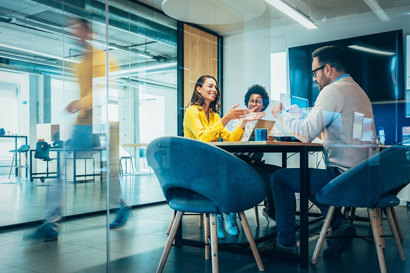 Three coworkers sit in a room with glass windows and have a meeting