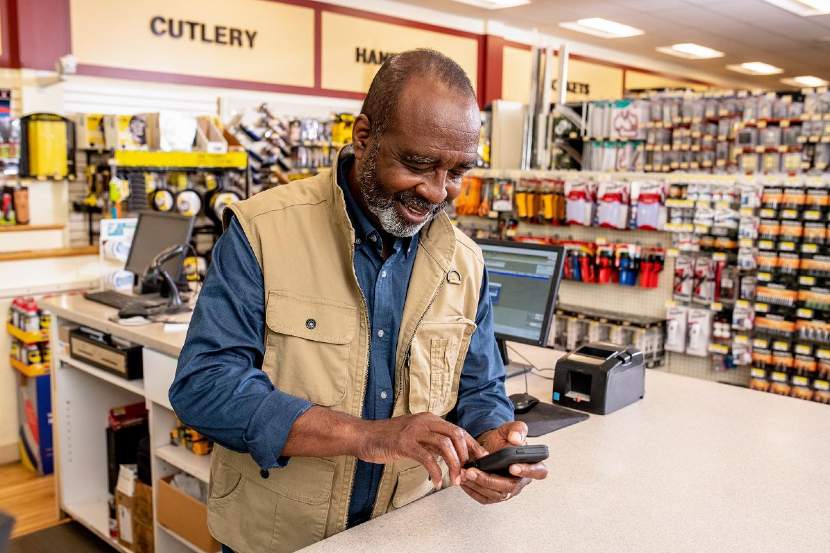 a man reviews his business metrics on his mobile phone while standing behind the register at his outdoor sporting goods shop