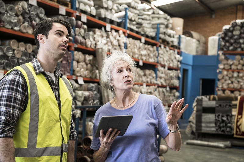 A warehouse manager stands in her warehouse holding a tablet and reviews inventory with an employee