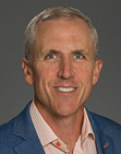 Dan Field has a short cropped grey hair, wears an orange dress shirt and blue suit jacket, and smiles for the camera