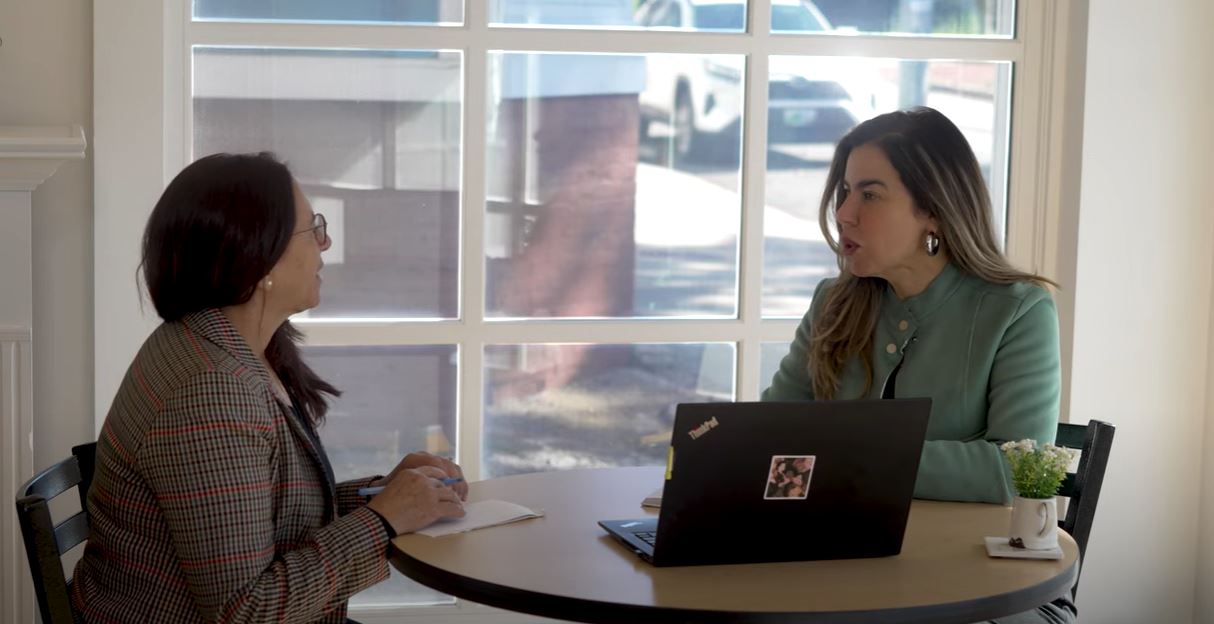 Two women sit at a table with a laptop and discuss business objectives