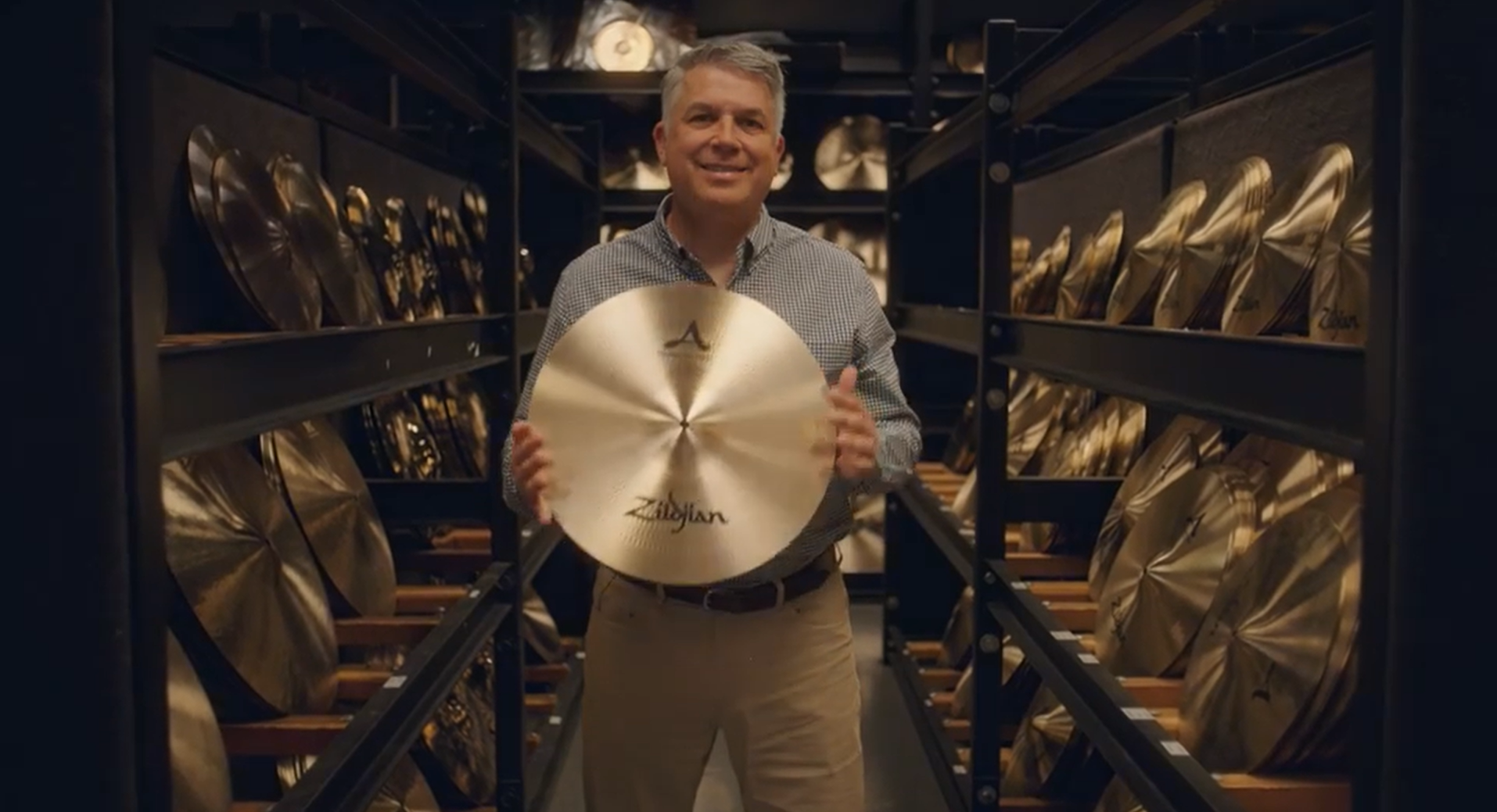 The CEO of Zildjian stands among racks of cymbals and proudly holds one in front of him for the camera