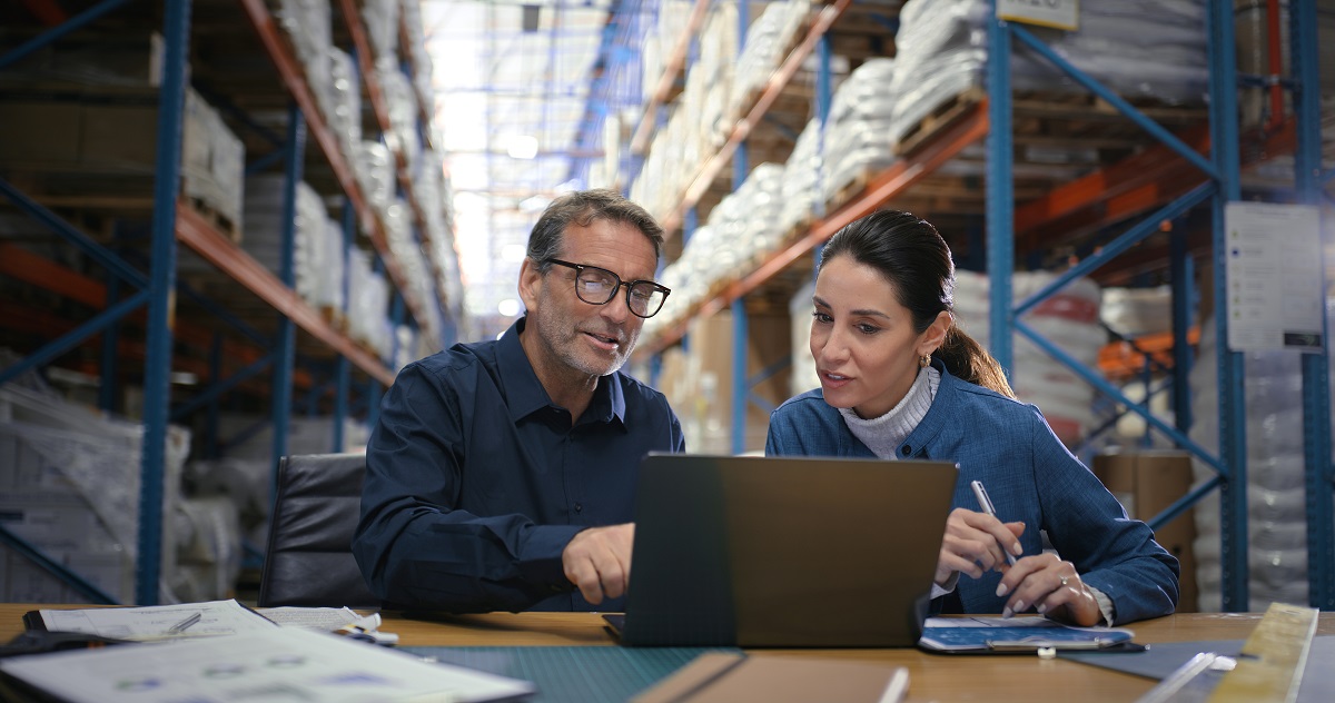 Two business partners sit together at a desk in a warehouse and review finances on a laptop