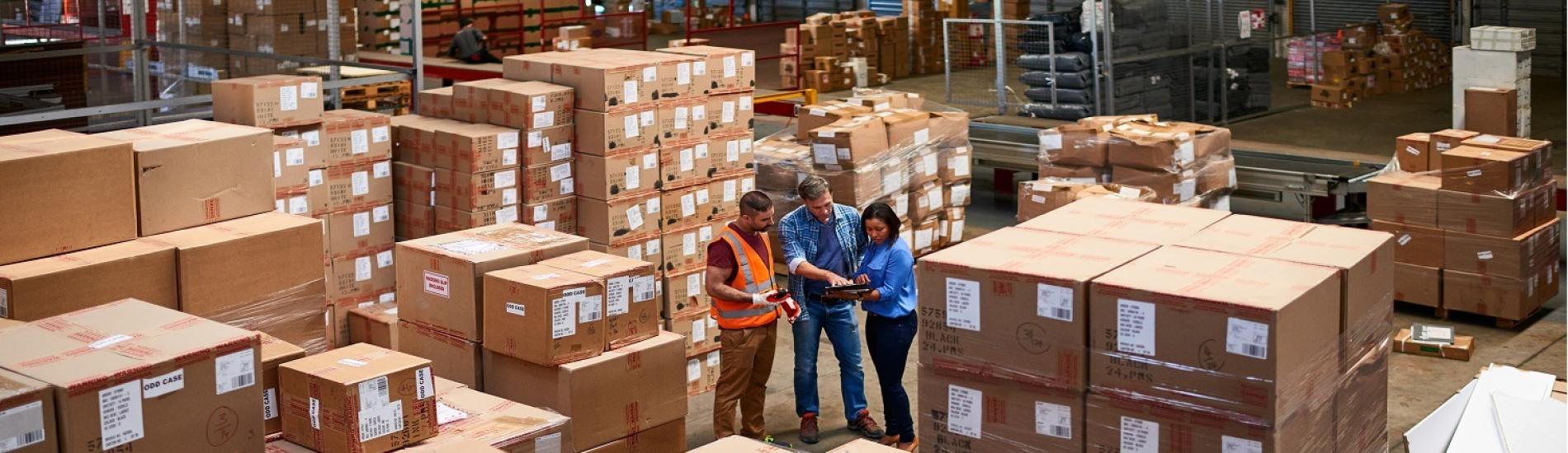 Three people stand in a warehouse amongst stacks of boxes and review business information on a tablet