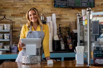 A woman stands at the checkout counter of her bakery while holding a computer tablet and smiling at the camera