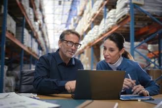 Two business partners sit together at a desk in a warehouse and review finances on a laptop