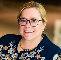 Nancy wears a dark blue blouse with flowers on it, wears black-rimmed glasses, and smiles at the camera