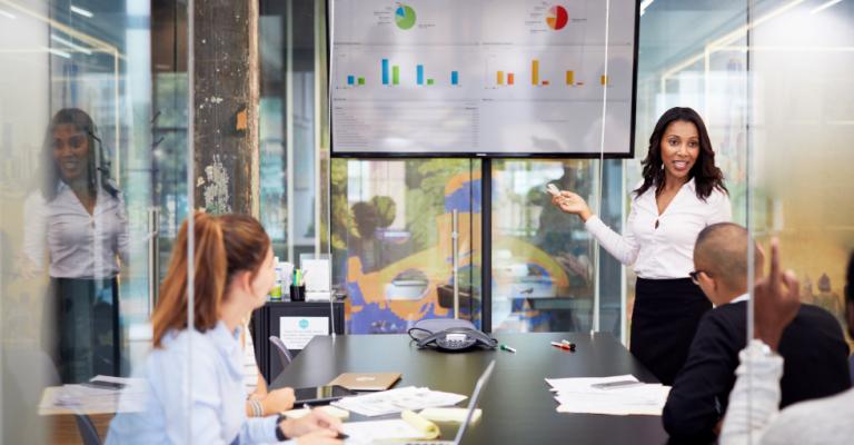 A woman stands in front of a screen displaying data and discusses business with colleagues in a meeting room