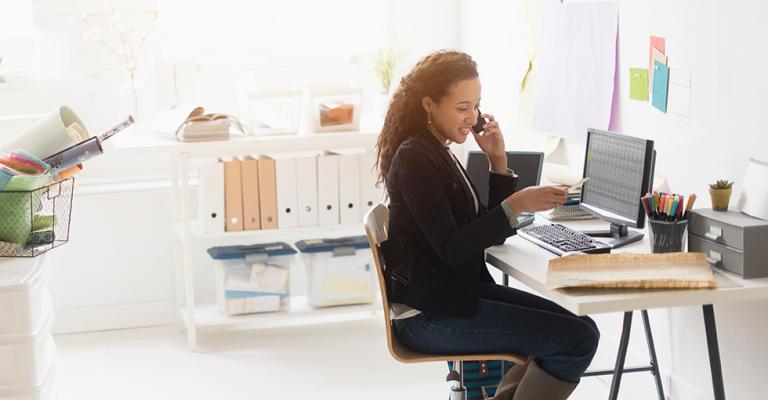 A woman sits at her desk reviewing finances on her laptop while speaking with her advisor on the phone