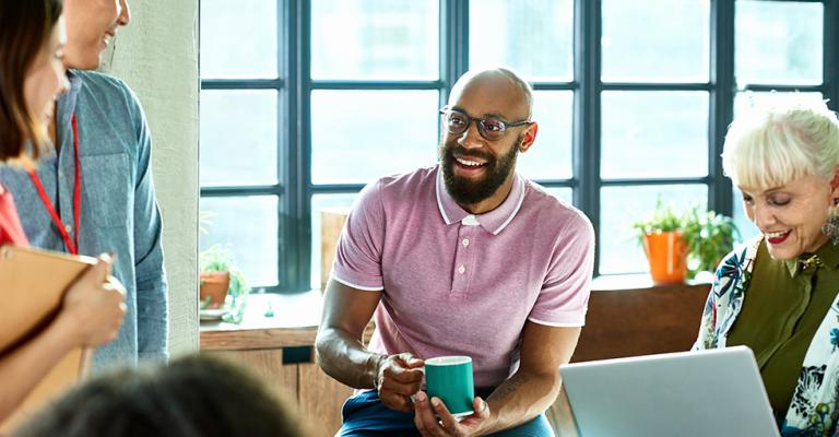 A man sits holding a cup of coffee and discusses business with his colleagues sitting around him