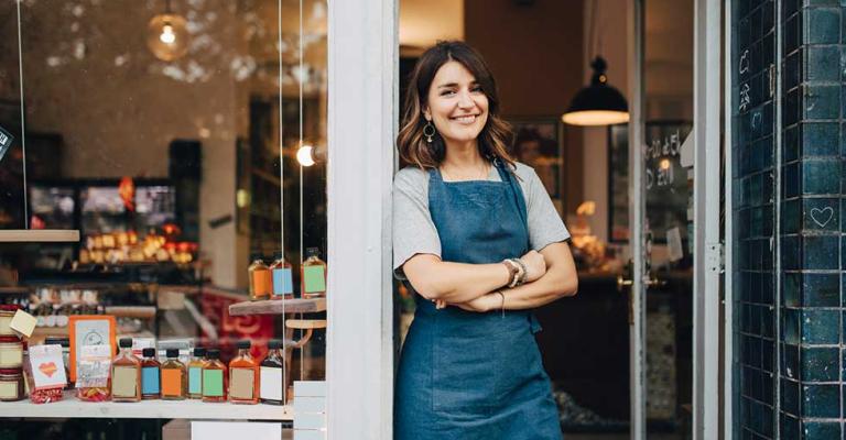 A person wearing an apron stands in the doorway of their bakery and smiles