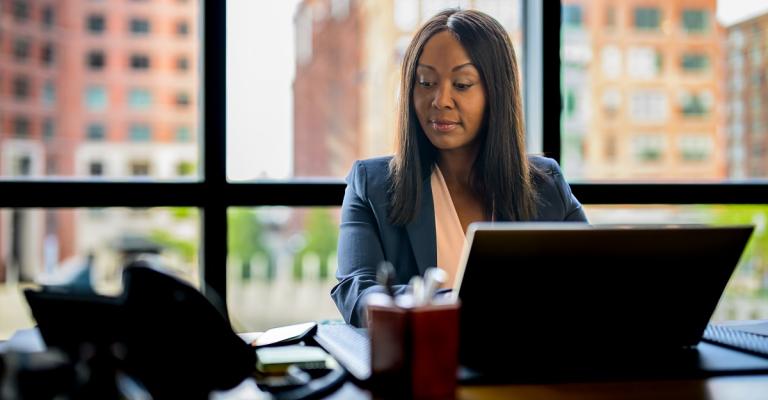 A woman sits at her desk and receives a fraud alert on her phone that she reviews on her laptop