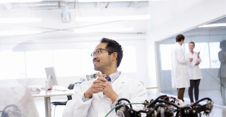 A scientist wearing a lab coat sits at his desk in his engineering lab reviewing his account on his computer