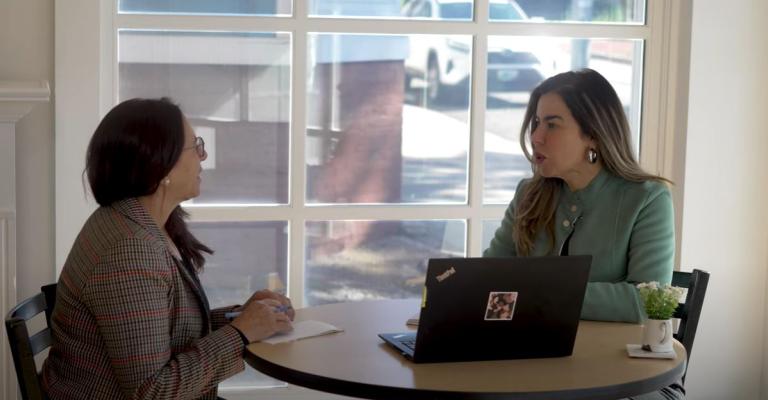 Two women sit at a table with a laptop and discuss business objectives