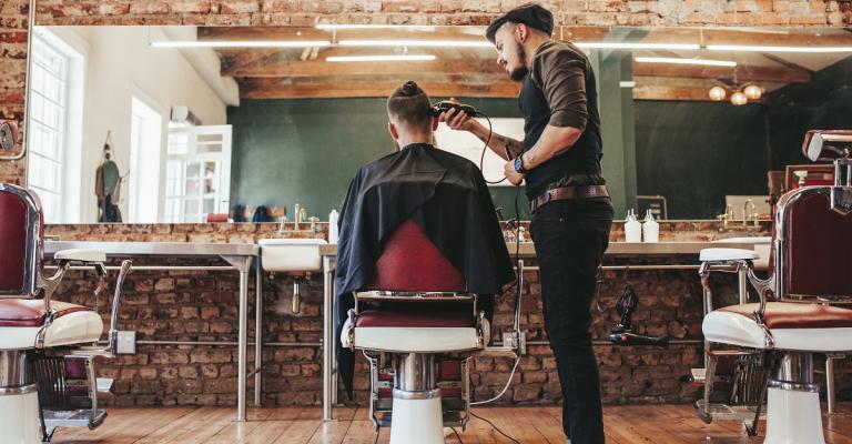 A man sits in a barber's seat and receives a haircut from the barber