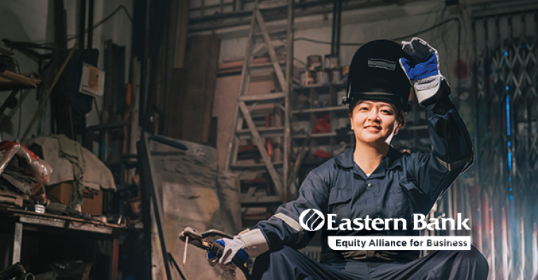 A woman sits in her metal shop and lifts her protective face shield to smile at the camera