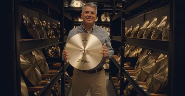 The CEO of Zildjian stands among racks of cymbals and proudly holds one in front of him for the camera